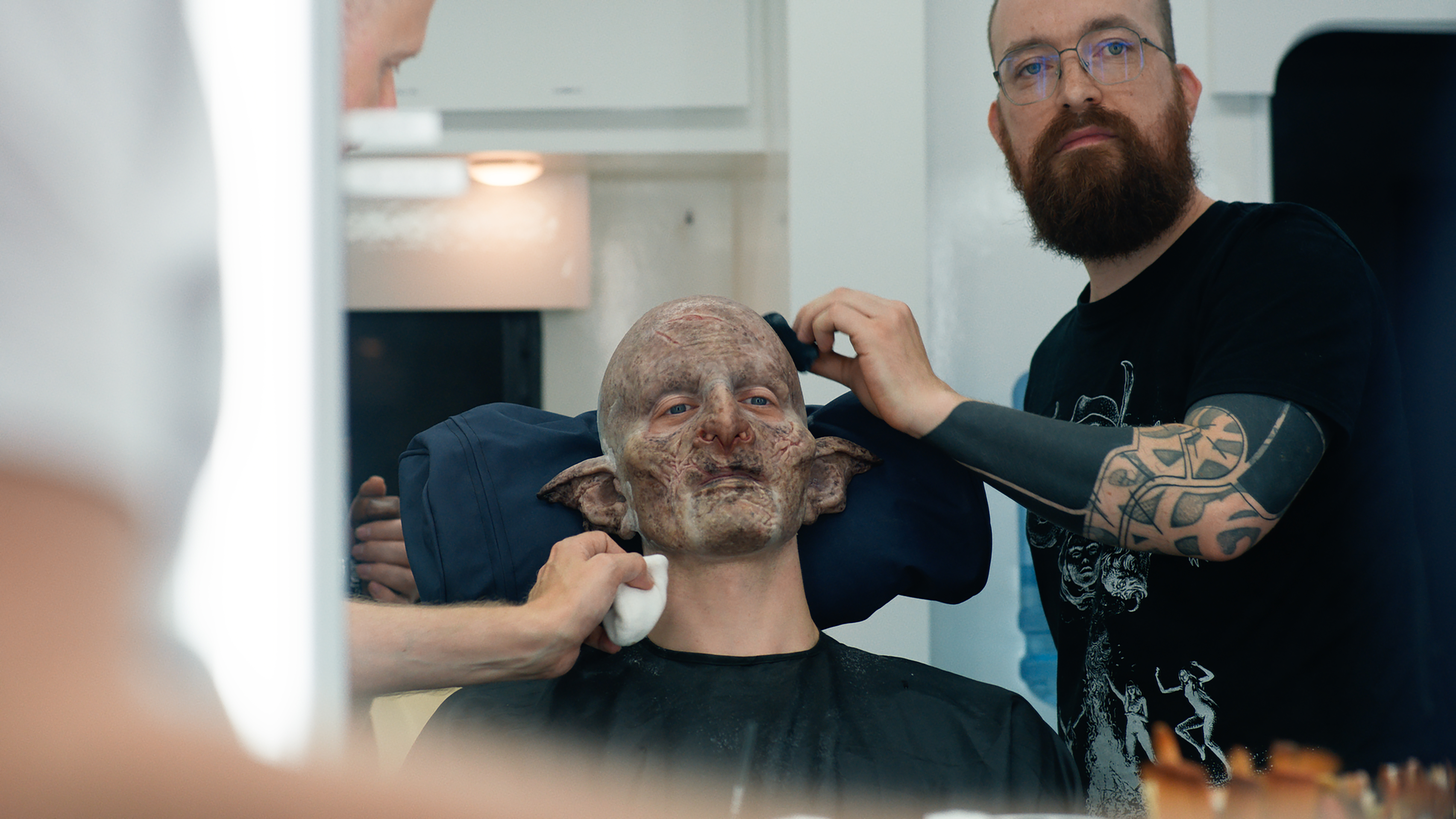 Robert Strange sitting in a chair in partial orc make-up with prosthetics artist Patt Foad tending to something on his head