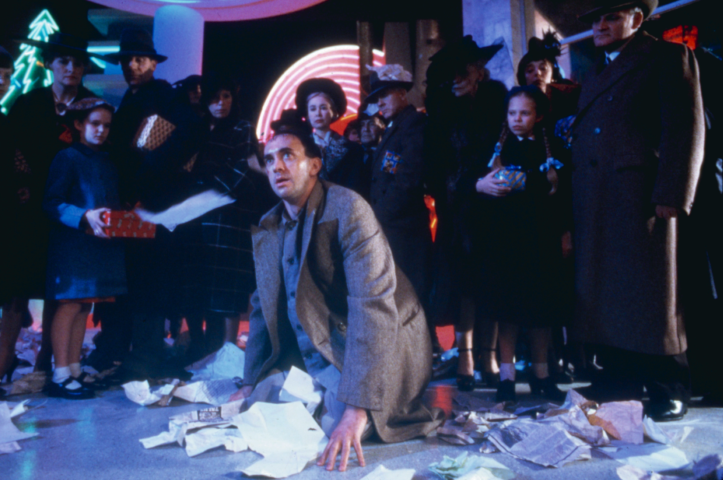 Sam Lowry (Jonathan Pryce) kneels amid a pile of scattered papers, surrounded by disapproving-looking, oppressively dressed people in a scene from Terry Gilliam’s Brazil