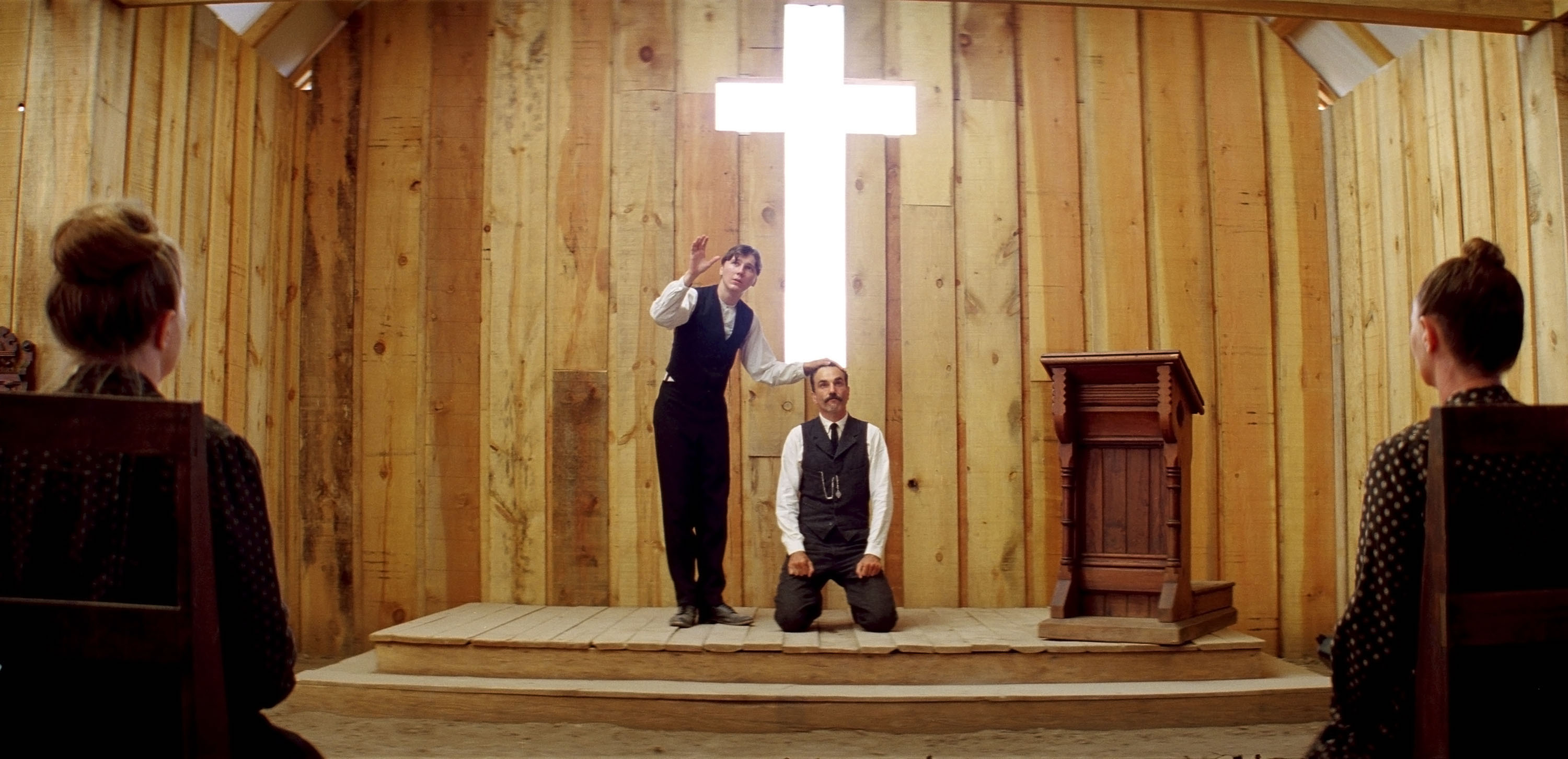 Daniel Plainview (Daniel Day-Lewis, in vest and shirtsleeves) kneels on a church dais with an immense glowing white cross behind him as Eli Sunday (Paul Dano) stands next to him, one hand on his head, the other raised in blessing. An audience watches them both. From Paul Thomas Anderson’s There Will Be Blood.