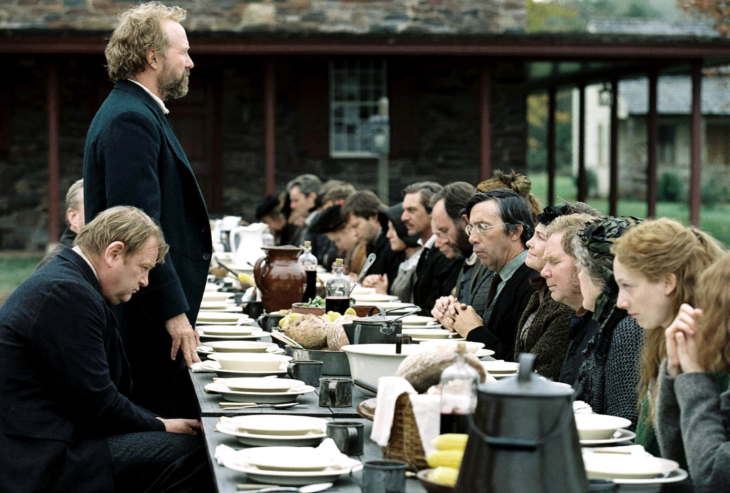 Several people sit at a table wearing early American clothing getting ready for a feast in The Village