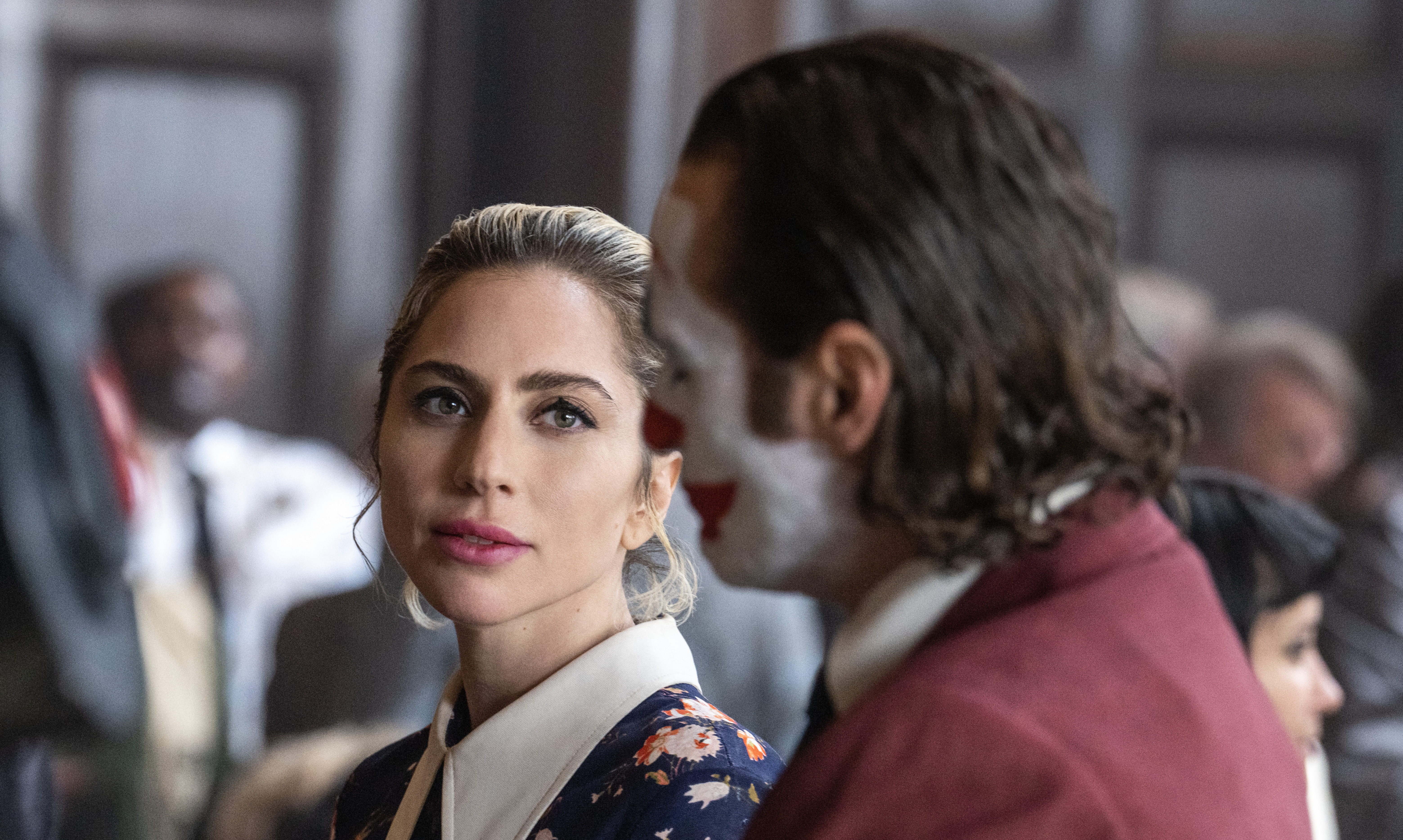 Harleen Quinzel (Lady Gaga) stands in a courtroom looking seriously at Arthur Fleck (Joaquin Phoenix, in full Joker regalia) in Joker: Folie à Deux