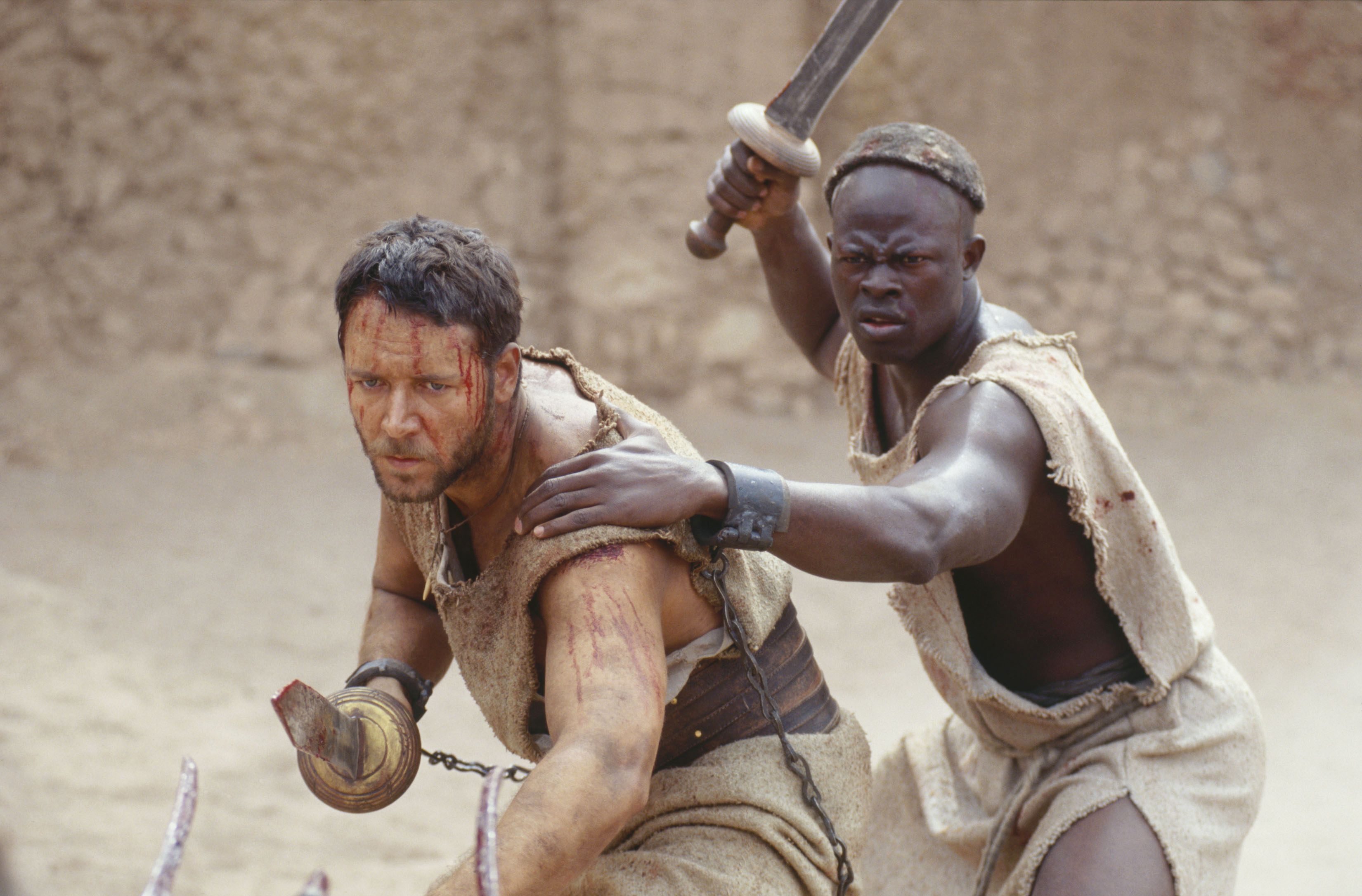 A pair of bloodied, dusty gladiators (Russell Crowe, Djimon Hounsou) stand together in the Colosseum, swords at the ready, in 2000’s Gladiator