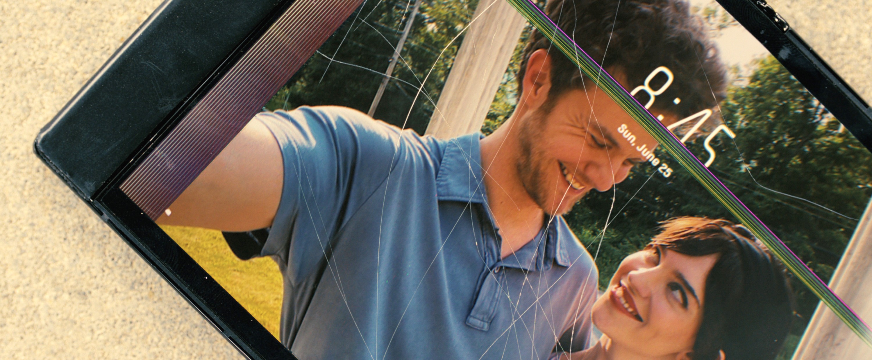 A cracked tablet screen shows an artifact-covered photo of young couple Iris (Sophie Thatcher) and Josh (Jack Quaid) smiling at each other in Companion