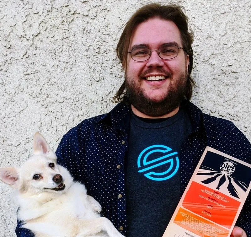 A photograph of Michael Clark, a smiling man holding a small white dog in one arm and a brochure for the Dice Awards in the other hand