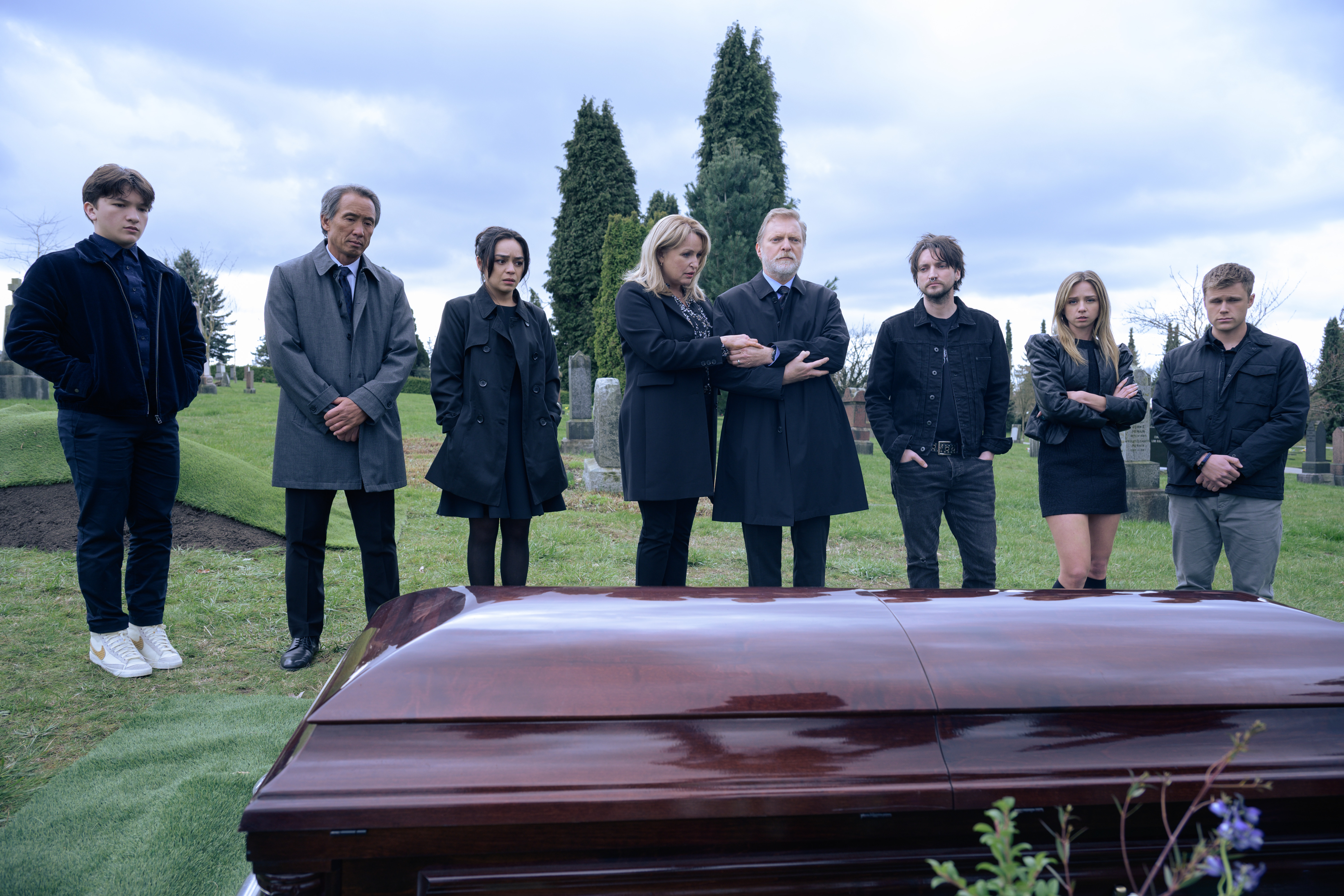 A line of characters in mourning black stand outside around a coffin in the foreground in Final Destinations: Bloodlines