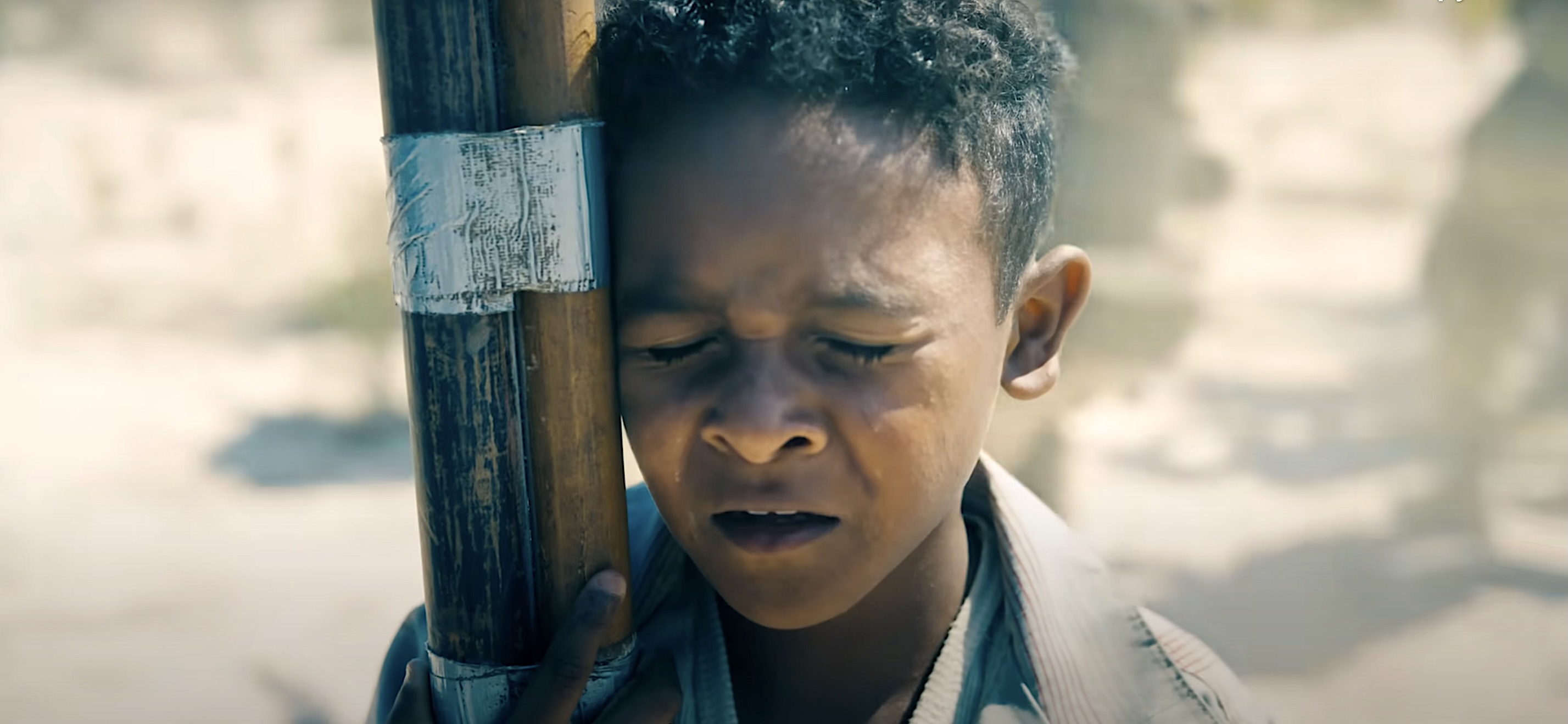 A thin, dirt-smeared, short-haired kid grimaces as he leans against a makeshift flagpole in the middle of a battleground in James Gunn’s Superman